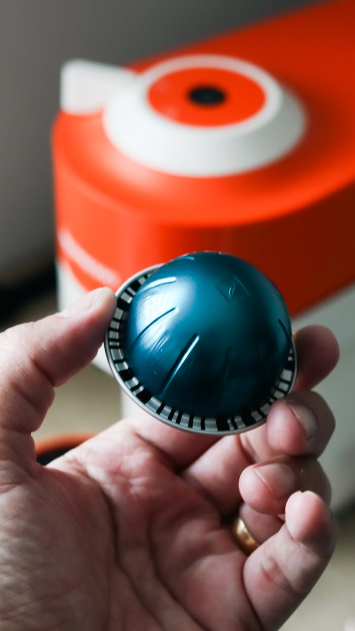 Close-up of a hand holding a coffee capsule in front of an orange coffee machine indoors.