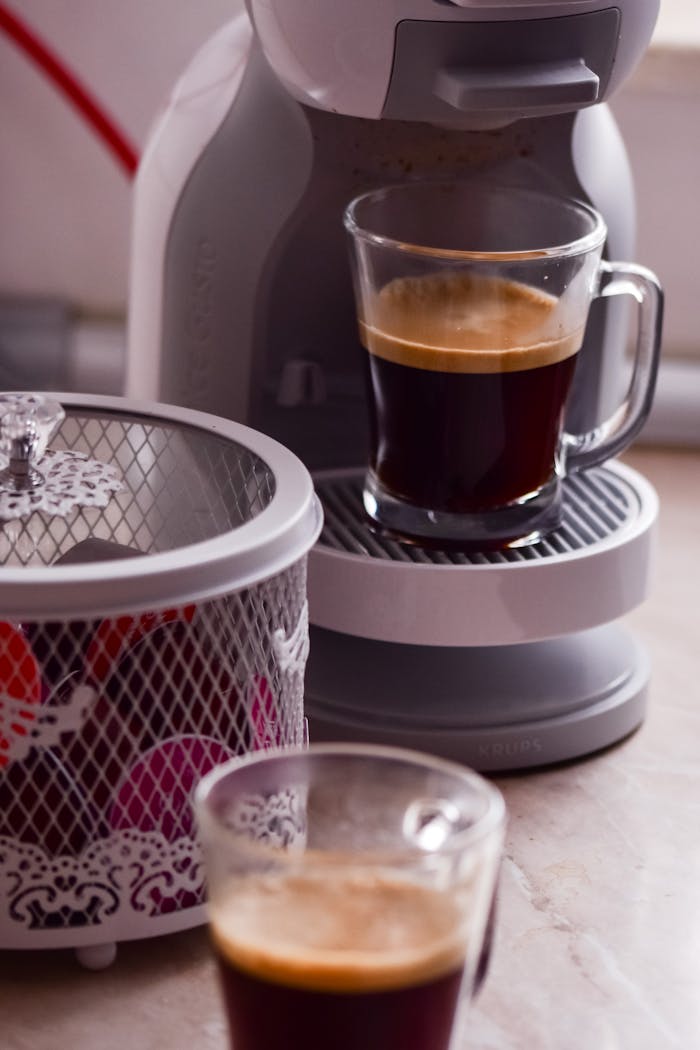 Modern professional coffee machine pouring fresh coffee into glass cup placed on wooden table near capsules in light kitchen at home